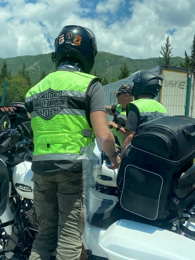 The image shows three individuals wearing motorcycle helmets and reflective vests, suggesting they are part of a motorcycle group. One individual, seen from the back, wears a black helmet with several patches and a bright green vest with the Harley-Davidson Cycles logo. Another individual, to the right, also wears a black helmet and a similar green vest. A third individual, partially visible between the first two, wears a camouflage cap and sunglasses, and is also in a green vest. They are standing next to white Harley-Davidson motorcycles. The environment appears to be outdoors, with a fence and a trailer visible in the background. Lush green mountains and trees suggest a rural or mountainous setting, possibly near Palje Brdo, Croatia, given the provided location context. The sky is partly cloudy, indicating daylight.