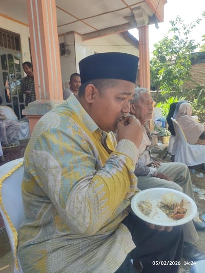 A man wearing a black peci and a yellow patterned shirt is eating from a white plate. He sits on a white chair with a yellow trim. Next to him sits an older man in a light-colored shirt. Behind them are people sitting in chairs, some wearing head coverings. The scene appears to be outdoors, possibly at an event or gathering, with a house in the background. The date "05/02/2026" and time "14:24" are stamped on the bottom right corner of the image.