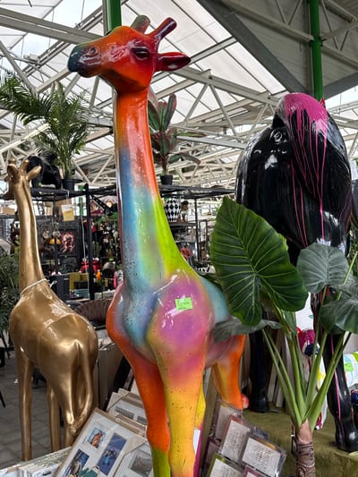 The image, taken in a retail environment in Aubange, Belgium, features several giraffe sculptures. The sculptures are in various colors and finishes: one is orange, yellow, and blue, another is gold, and a third is black with pink streaks. The giraffes are arranged near each other, with other retail items visible in the background on shelves and displays. The environment is illuminated by natural light entering through a glass roof, which is supported by a metal framework.