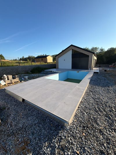The image depicts an outdoor scene in Horní Planá, Czechia, featuring a newly constructed swimming pool area next to a modern house. The swimming pool is rectangular with light blue lining and a shallow green section, surrounded by a tiled patio. The patio is bordered by a metal edge and gravel. To the right, a contemporary house with a distinctive roofline, white and wooden facade, and dark metallic shutters is visible. The surrounding environment is characterized by a clear blue sky, trees in the background, and a fence along the property line. Construction materials, including concrete blocks and pallets, are scattered in the foreground and to the left, indicating ongoing development. The ground is covered with gravel and dirt, and there is evidence of unfinished landscaping.