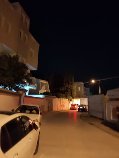 At night in an urban setting, a white car is parked in the foreground. Beyond it, a street curves to the right, with a single streetlamp illuminating the scene. Two cars, a red car and a dark-colored car, are driving away from the viewer on the street. Residential buildings are visible on the left, and a fence lines the right side of the street. A person stands on the right side of the street.