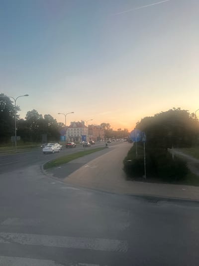 The image shows a wide street in Lublin, Poland, during sunset. The sky is a gradient of blue and orange, with a faint contrail from an airplane visible. On the left side of the street, there are trees and streetlights. Several cars are driving on the road, including a white sedan and a black SUV. In the middle of the road, there is a pedestrian crossing. On the right side of the street, there is a sidewalk with a row of trees and bushes. Traffic signs are visible, including a pedestrian crossing sign and a bicycle lane sign. In the background, there are buildings with traditional architecture. The overall scene depicts a typical urban environment at dusk.