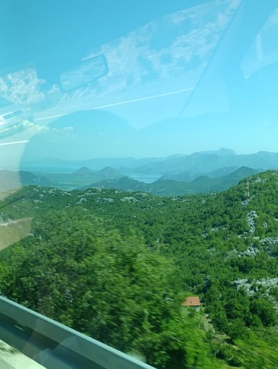 The image shows a panoramic view of a mountainous landscape with lush green vegetation. In the foreground, a blurry green tree dominates the left side, indicating movement, likely from a vehicle. A gray guardrail runs along the bottom edge of the frame. The middle ground features rolling hills covered in dense green trees, interspersed with patches of lighter-colored rocks. A small, orange-roofed building is nestled among the trees on the right side of the middle ground. In the background, a serene body of water is visible, surrounded by layers of hazy blue mountains under a clear sky with wispy white clouds. The overall scene suggests a scenic drive through a natural environment, possibly in the vicinity of Gornja Brova, Montenegro, given the characteristic mountainous terrain and vegetation. The image appears to be taken from inside a vehicle, evidenced by the reflections on the glass and the blurred foreground.