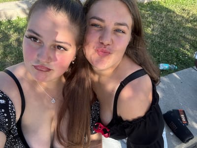 Two young women are posing for a selfie outdoors on a sunny day. The woman on the left has dark brown hair, fair skin, and is wearing a black and white floral top, a silver necklace, and has a nose ring. She is looking towards the camera with a slight pout. The woman on the right has light brown hair, fair skin, and is wearing a black off-the-shoulder top. She is also looking towards the camera with her lips puckered. They are positioned close together, with the woman on the right slightly behind and to the right of the woman on the left.

The background consists of green grass with some patches of brown and dry areas, suggesting it might be a park or an outdoor recreational area. A grey concrete surface runs along the bottom and right side of the image, possibly a path or seating area. A discarded plastic water bottle is visible on the grass in the middle ground. A black portable speaker with "JBL" branding and what appears to be a packet of rolling papers are placed on the concrete surface on the right side of the frame. The lighting indicates it is daytime, with shadows cast on the grass, suggesting the sun is at an angle.

While the exact location in Weil am Rhein, Germany cannot be definitively determined from the visual information alone, the presence of a water bottle and a portable speaker, along with the casual outdoor setting, might suggest a public park, a recreational area along the Rhine river, or an outdoor gathering spot within the city.