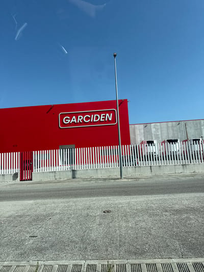 The image shows a bright, sunny day in Massanes, Spain, with a clear blue sky. The main subject is a large, red building with the white text "GARCIDEN" prominently displayed on its side in a vertical orientation. The building appears to be a commercial establishment, possibly a warehouse or retail store, with a modern design characterized by a white horizontal railing or fence running along its length.
Behind the red building, another section of the structure is visible, made of concrete panels and with multiple white fences or barriers. Several white trucks are parked in a line behind these barriers, suggesting a logistics or distribution operation is taking place. A street or road made of grey asphalt runs alongside the building and fence, with some textured concrete barriers on the far right. A lamppost is visible in the foreground, extending from the left side of the frame towards the red building.
The overall impression is one of a business or industrial setting on a clear day. There are no people visible in the image. The bright sunlight indicates it is daytime, likely mid-morning or afternoon, and the clear sky suggests fair weather. The text "GARCIDEN" is the only discernible text in the image.