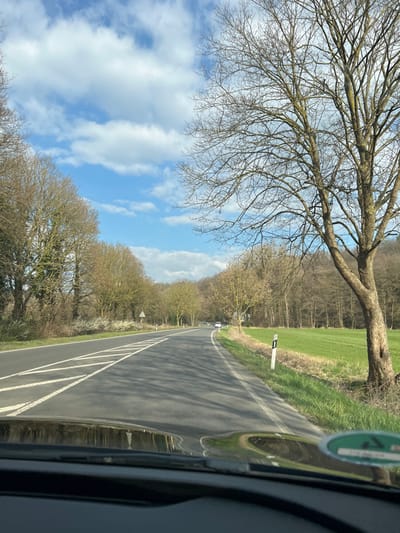 The image captures a forward-facing view from inside a vehicle on a paved road in Stolberg, Germany. The road, marked with white dashed lane lines and a solid white edge line, curves gently to the right in the mid-distance. Tree shadows are distinctly cast across the road surface.
To the left of the road, a dense line of deciduous trees, mostly bare, borders the route, with some exhibiting early spring foliage. A white triangular road sign with a red border is visible on the left shoulder. To the right of the road, a grassy verge leads to a vibrant green field, which is backed by a dense deciduous forest covering a sloping terrain. A prominent, large, bare deciduous tree with extensive branching stands on the right side of the road, near the verge. A white roadside marker post with a black top is situated on this verge.
A white car is distantly visible moving away on the curved section of the road. The sky above is blue with scattered white cumulus clouds. The lower part of the image shows the vehicle's dark dashboard and a reflection of a circular green environmental sticker (Umweltplakette) on the windshield. No individuals are visible.