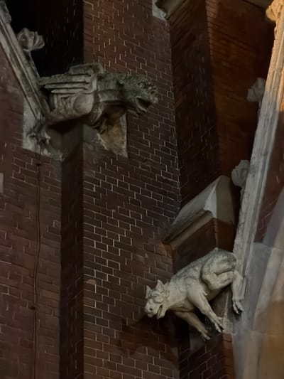 This image shows two gargoyle statues attached to a brick wall or roof structure. The statue on the left is a winged figure, possibly a griffin or a similar mythological creature. The statue on the right depicts an animal, perhaps a dog or a boar, hanging upside down. The architecture suggests a historical building. These gargoyles are on the main post office in Osijek, Croatia.