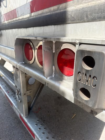 The image shows the rear of a silver trailer, likely a semi-trailer, in what appears to be a commercial or industrial area. The trailer has red and white reflective tape along its lower edge and a black rubber rub rail above the taillight assembly. Three round red LED taillights are visible, recessed into a metal housing. One of the lights has "TOP" and "Grote" branding. To the right of the taillights is a rectangular metal housing with two oval cutouts, one of which displays the embossed letters "CIMC". The ground is asphalt, with some debris visible. The overall impression is a functional, utilitarian vehicle component. The scene's specific location within the city of Lebanon, United States of America, is not discernible from the image alone.