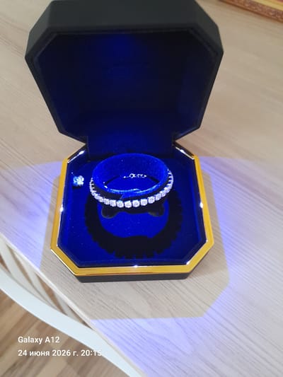 The image shows an opened jewelry box, which is black and hexagonal with a gold trim. Inside, the box is lined with dark blue velvet and contains a sparkling diamond bracelet arranged in a circular fashion on a velvet stand. To the left of the bracelet, a small, single diamond is visible. The jewelry box is placed on a light-colored wooden surface. The lower left corner of the image displays a timestamp and device information: "Galaxy A12" and "24 июня 2026 г. 20:15". The context of Kara-Köl City, Kyrgyzstan is not depicted in the image.