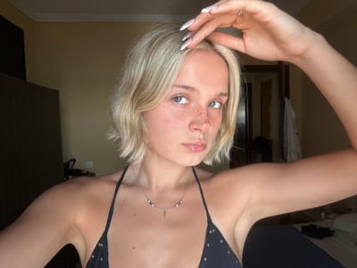 This is a close-up, upside-down portrait of a young Caucasian woman with blonde hair. She is wearing a black bikini top adorned with metal studs and a delicate silver necklace with small beads. Her face is turned towards the viewer, and she has striking green eyes and a smattering of freckles across her nose and cheeks. Her lips are slightly parted, and she has a neutral expression. Her left arm is raised, and her hand is positioned near her face, with her fingers delicately touching her hair. Her nails are long and painted with a white and silver design. The background is softly blurred, showing a wall and what appears to be furniture, suggesting an indoor setting. The lighting is warm and even, indicating it is likely daytime or well-lit indoors. No visible text is present in the image.