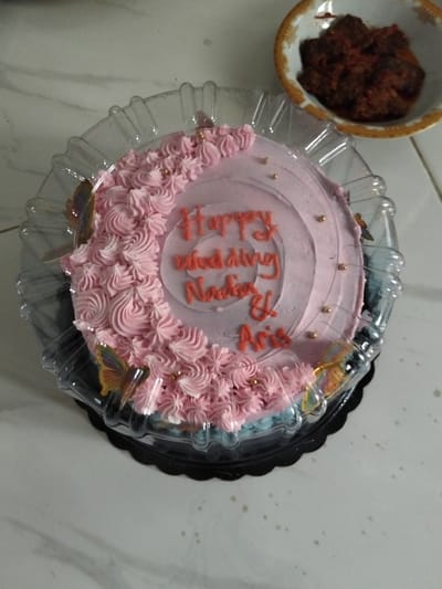 A pink frosted cake sits in a clear plastic container on a marble-patterned surface. The cake is decorated with piped pink frosting around the edge and has "Happy Wedding Nadia & Aris" written in orange icing in the center. Several edible gold balls are scattered on the cake. Two decorative butterflies, one orange and one iridescent, are attached to the side of the cake. A small bowl of what appears to be food is visible in the upper right corner of the image. The scene is indoors, suggesting it might be a home or a bakery in Cottrueng, Indonesia.