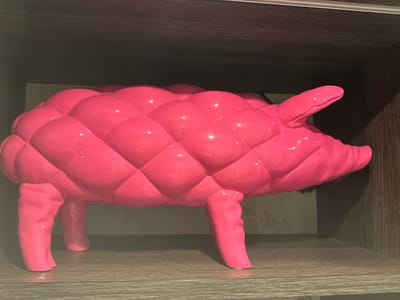 The image shows a bright pink, quilted-pattern pig figurine. It has a plump body with a pattern of raised, rounded squares resembling a quilted texture. The pig has short, stubby legs, a curled tail, and a protruding snout. It is placed on a shelf with a light-colored, wood-grain finish. Behind the pig, there is a striped pattern, suggesting a background of some kind. The lighting is even, highlighting the glossy finish of the figurine. The setting is simple and lacks any distinguishing features that would connect it to Genk, Belgium or any other specific location.