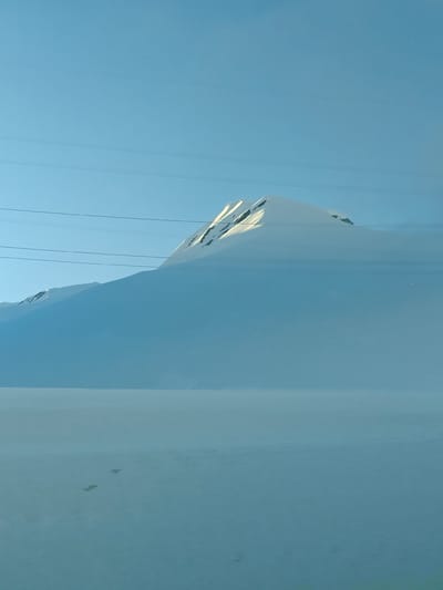 This image captures a vast, serene, snow-covered mountainous landscape in Ganisi, Georgia, under a clear, bright blue sky.
The prominent subject is a snow-capped mountain that dominates the upper and mid-ground. Its peak is brightly illuminated by direct sunlight, highlighting the pristine white snow and rocky outcrops. The lower slopes of the mountain appear partially obscured by shadows or a soft atmospheric haze, blending into the expansive, flat snowfield that stretches across the foreground and mid-ground. This snowfield is largely unbroken, creating a sense of immense scale and tranquility, with a few indistinct dark specks visible on its surface in the lower half of the frame, possibly small rocks or distant objects.
Several thin, dark utility lines traverse horizontally across the sky and mountain, introducing a man-made element into the otherwise natural scene. The sky is a uniform, vivid blue, suggesting a clear day. A faint, slender crescent shape, likely the moon, is visible high in the sky towards the upper right of the frame.
The time of day appears to be either morning or late afternoon, indicated by the angle of the sun brightly illuminating the mountain peak while leaving other areas in shadow. The weather is clear and sunny, implying cold temperatures typical of a snowy mountain environment. There is no visible text in the image. The overall impression is one of cold, quiet grandeur and expansive natural beauty.