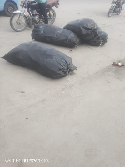 In Adesanya, Nigeria, three large black trash bags lie on a concrete surface, one in the foreground and two further back. The bags are tied with white string, with one bag in the foreground clearly displaying text. A motorcycle, with a person sitting on it, is visible in the background to the left. The motorcycle appears to be a common model in the region. To the right of the trash bags, a brown plastic bottle lies on the ground. The scene is outdoors, likely during the day given the lighting. The ground appears to be dusty and worn, suggesting a public area or roadside. The overall impression is one of everyday life and perhaps waste disposal in a bustling environment. The visible text on the trash bag appears to read "SA". The overall scene is mundane and representative of daily occurrences in the specified location.