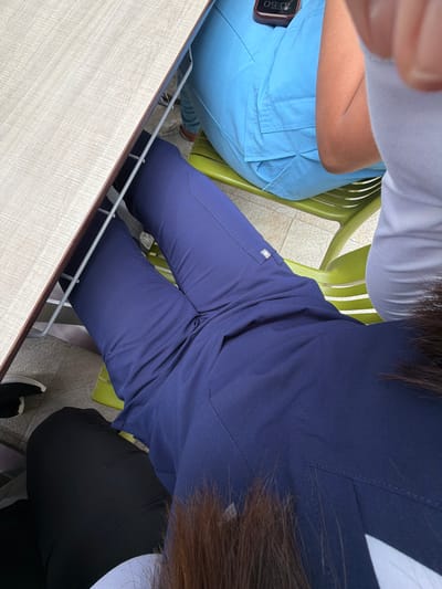 An overhead shot captures three individuals seated around a table. The central figure wears dark blue scrubs, their dark hair partially obscuring their back. A small, white, rectangular logo is visible on the right thigh pocket of their scrubs. They are seated on a bright green plastic chair. To their left, another person is seen wearing light blue scrubs with pleated detailing on the back. A mobile phone with the time "10:50" displayed on its screen is tucked into a cargo pocket on their scrubs. This individual is also seated on a matching green plastic chair. A third person is partially visible in the bottom right, wearing black pants and black-and-white athletic shoes. A light-colored wooden table with a metal frame forms the foreground of the image. The setting appears to be a casual indoor or covered outdoor area, possibly a break room or cafeteria, given the attire and furniture.