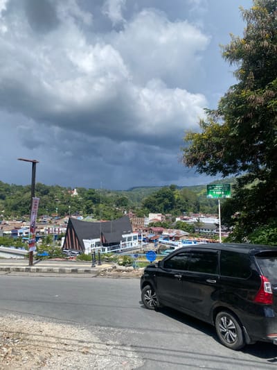 A black minivan is parked on a paved road overlooking a town nestled in a valley, with lush green hills and a dramatic, cloudy sky in the background. The town features a distinctive building with a black, angled roof and white walls, likely a public building or church. Other buildings in the town are of various colors and architectural styles. A green sign with white lettering is visible on the right side of the road, indicating "PELABUHAN FERRY" (Ferry Port) and a distance of 950 meters. A lamppost with a banner hangs on the left side of the road. The environment appears to be a scenic overlook in Parapat, Indonesia, possibly near Lake Toba, given the ferry port sign. The scene is devoid of people.