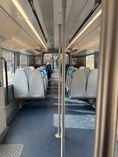 The image shows the interior of a modern commuter train carriage, stationary at a platform within the Elmbridge Borough, United Kingdom. The carriage features a central aisle flanked by two rows of forward-facing seats, which are light grey with blue accents on their upper sections. The floor is blue with a speckled pattern. Overhead, the ceiling is white with linear strip lighting extending the length of the carriage. Several silver-colored vertical poles, designed for standing passengers, are visible, one prominent in the right foreground.
Through the windows on the left side, a train platform is visible, with a male individual standing on it. He is dressed in a dark jacket and positioned with his back partially towards the camera, near the train's doorway. A second, less distinct figure is further down the platform. Beyond the platform, trees and outdoor foliage are discernible. Through the windows on the right, an exterior wall or embankment with a textured, possibly stone or concrete, surface is visible.
Within the train, additional passengers are partially visible further down the aisle. An overhead CCTV camera is mounted at the far end of the carriage. Information posters are affixed to the internal walls. A green illuminated button, part of the door control mechanism, is present on the left door frame. The scene is lit by interior lighting and ambient daylight entering through the windows.