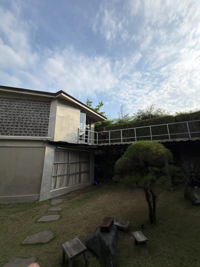 This image depicts an outdoor scene in Tumbak Bayuh, Indonesia, featuring a modern architectural structure and a landscaped garden under a partly cloudy sky.
The primary subject is a two-story building constructed with grey concrete and a section of textured grey brickwork on its upper facade. A dark roof covers the structure. On the upper level, a glass door opens onto an elevated, flat walkway or bridge with grey metal railings. One individual, dressed in dark attire, is standing on this walkway, facing left. The lower level of the building features a wall with large, grid-framed windows, behind which light-colored curtains are visible.
The ground area in front of the building is a green lawn. A winding path of irregular grey stone stepping stones traverses the grass towards the building. In the foreground, several rustic outdoor furniture pieces are present: two square wooden stools and a larger dark, irregular base (possibly stone or concrete) topped with a rectangular wooden box. A small, gnarled tree with green foliage stands on the right side of the lawn. Beyond the elevated walkway, a dense hedge of green foliage forms a boundary, with taller trees and bamboo visible above it. On the far right, partially obscured by vegetation, a second individual is visible, seated or crouching on the grass, wearing dark clothing.