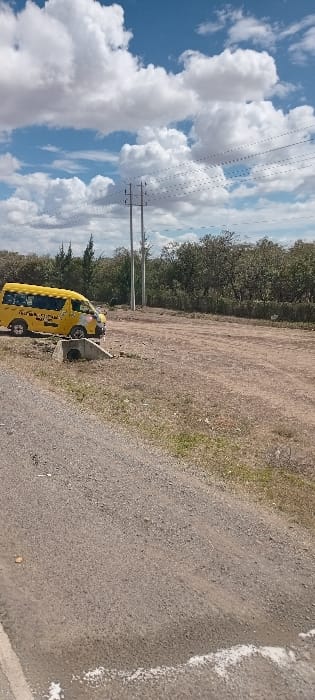 A yellow van is parked along a gravel road. Adjacent to the road is an open dirt area with some sparse grass growth. Beyond that, there is a row of trees. The sky is visible, filled with numerous white clouds and power lines. The image has a slight tilt.