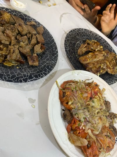 Three plates of food are displayed on a white tablecloth. The first plate, with a black and white patterned design, holds grilled lamb chops. Another matching plate contains cubed grilled meat. The third plate, white, features grilled meat topped with sauteed onions and sliced tomatoes. A small container of sauce is visible near the cubed meat plate. Human hands are visible at the top left of the image.
