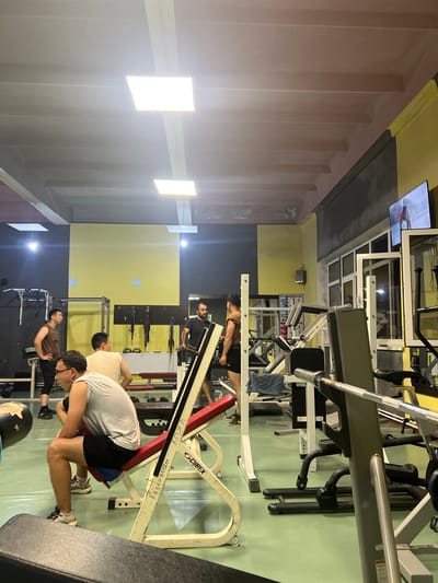 The image depicts the interior of a gym, likely located in Kadamjay, Kyrgyzstan. Several individuals are present, engaged in various activities. In the foreground, a man wearing a gray sleeveless shirt and black shorts is seated on a red and white weight bench, looking to his left. To his left, a portion of a large, dark, spherical object is visible. Behind him, another man in a white tank top is also seated. To the left of the bench, a man in a brown tank top and black shorts stands with his hands on his hips, facing away from the camera. Further back, another man in a dark shirt stands near a weight rack. In the background, a television screen is mounted on the wall, displaying an image of a person. The gym environment includes various weightlifting equipment, such as benches, racks, and barbells. The walls are painted in a combination of yellow and dark gray, with fluorescent lighting fixtures on the ceiling. The floor is a light green color.