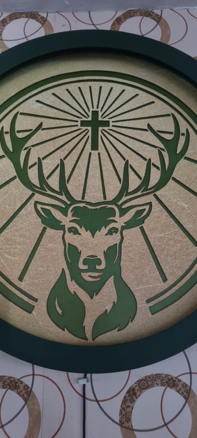 The image shows a circular Jägermeister logo hanging on a wall. The logo features a green deer head with antlers inside a wheel, with a Christian cross positioned above the deer's head. The background of the logo is a textured gold color, and the outer ring is dark green. The wall behind the logo has a patterned wallpaper with circles. The location is unknown; there is no information to indicate the scene is in Alasia, Nigeria.