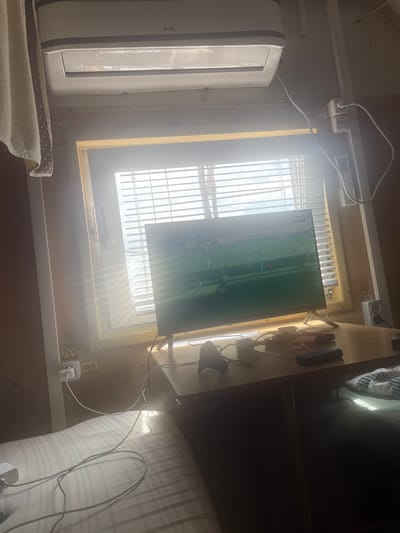 The image depicts an indoor scene, likely a small room or living space, with bright sunlight streaming through a closed window covered by horizontal blinds. A modern flat-screen television is mounted on a stand on a wooden desk, displaying a soccer match. On the desk, there are various electronic accessories, including chargers, cables, and a dark-colored rectangular object, possibly a power bank or external hard drive. To the left of the desk, a white air conditioning unit is mounted high on the wall. A towel with a polka dot pattern hangs from the left side of the frame. Electrical outlets and warning labels are visible on the wall to the right of the window. The foreground shows a portion of a bed with a striped mattress protector. The overall impression is of a functional, albeit somewhat cluttered, personal space. The location is stated to be Tatishchevo, Russia.