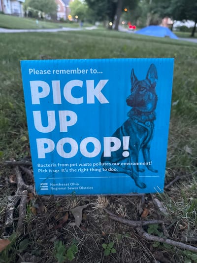 A blue corrugated plastic sign with white text and an image of a dog is placed in a grassy area with fallen branches and leaves. The sign reads, "Please remember to... PICK UP POOP! Bacteria from pet waste pollutes our environment! Pick it up. It's the right thing to doo." Below this text is a logo that says "Northeast Ohio Regional Sewer District." The background shows a blurred view of houses, trees, and a blue tarp on the ground, suggesting a residential or park area within Shaker Heights, Ohio, United States of America. The lighting indicates it is daytime, possibly late afternoon or early evening.