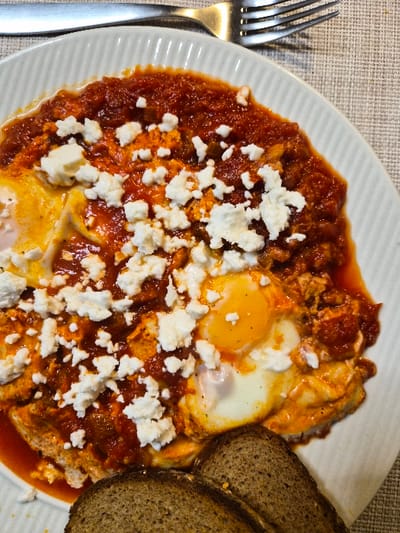 A high-angle, close-up photograph displays a meal served on an oval white ceramic plate with a ridged rim, situated on a textured, light beige woven tablecloth. The main dish consists of at least three visible cooked eggs with intact yellow-orange yolks and off-white whites, submerged in a thick, chunky, reddish-orange tomato-based sauce. Numerous irregular white crumbles of feta cheese are scattered liberally over the entire surface of the sauce and eggs. Two slices of dark, whole-grain bread are positioned at the bottom right of the plate, partially covered by the sauce. A silver-colored metal fork with four tines is visible in the upper right corner, resting on the tablecloth. The scene is located in the Municipal Unit of Corinth, Greece.