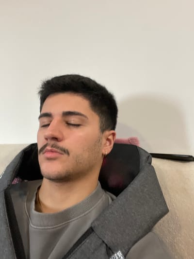 The image depicts a young adult male in São Mamede de Infesta, Portugal, seated indoors, with his eyes closed. He has short dark hair, a mustache, and light stubble. A small gold hoop earring is visible in his right ear. He is wearing a plain grey crew-neck sweatshirt and utilizing a grey neck and shoulder massage device which extends over his shoulders, with a black mesh section visible at the neck. He appears to be resting or relaxing. Behind him, a light-colored textured surface, likely a sofa or chair, is partially visible. A small fluffy pink object and a dark rectangular object (possibly a smartphone or remote control) rest on this surface behind the massage device. The background consists of a plain, light-colored wall.