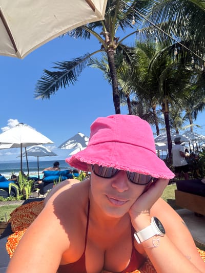 A selfie captures a woman in a pink hat and sunglasses on a sunny day at a beach in Medahan, Indonesia. She is wearing a white smartwatch with a heart bracelet on her wrist and a red swimsuit. Behind her, the clear blue sky is dotted with white beach umbrellas. Palm trees frame the scene. In the background, people are visible on lounge chairs near the ocean. A white umbrella and vegetation are also present.
