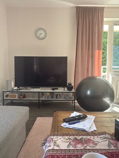 The image depicts a living room interior, likely in a residential setting. Prominently featured is a large, dark-colored television screen positioned on a white marble-topped media console with black metal legs. To the left of the TV, a grey cylindrical speaker is visible. A round, grey exercise ball is situated to the right of the media console. In the background, a floor-to-ceiling pink curtain hangs by a window, through which greenery and sunlight are visible. A round wall clock with black numbers and hands is mounted on the wall above the television. On a wooden coffee table in the foreground, a stack of papers, a black remote control, and a blue pen are arranged. A part of a grey upholstered sofa is visible on the left, and a light pink rug is placed on the floor between the sofa and the coffee table. The overall scene suggests a casual and lived-in domestic environment. The presence of an exercise ball and papers with writing implies potential activities such as fitness and note-taking or studying. The lighting suggests it is daytime, with sunlight streaming in from the window. This scene is not identifiable as being in a specific city or district.