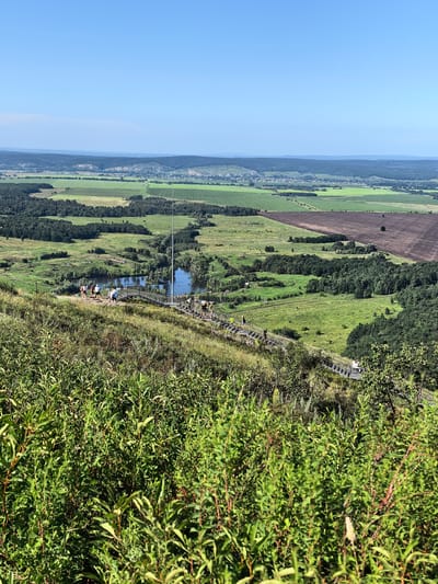 An aerial shot captures a panoramic view of the city of Shikhan, Russia, dominated by a vast, clear blue sky. In the foreground, lush green foliage partially obscures the view. A wooden walkway or staircase descends from the upper right to the lower left, with several individuals walking on it. Below the walkway, a tranquil body of water, likely a lake, is visible, reflecting the surrounding trees and sky. The landscape unfolds into rolling hills covered with green fields, trees, and a distant town or village nestled in a valley. The terrain suggests a natural, hilly landscape with agricultural land and forested areas, under a bright, sunny sky.
