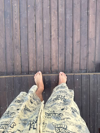 A first-person perspective shot shows a person's bare feet and patterned shorts standing on a wooden deck. The deck is made of dark brown planks running vertically, with some grass growing between the planks. The shorts have a light yellow base with black graffiti-style text and designs. One foot is positioned slightly in front of the other, with the toes of the front foot visible. The image is taken from a high angle looking down. The location is not specified in the image.