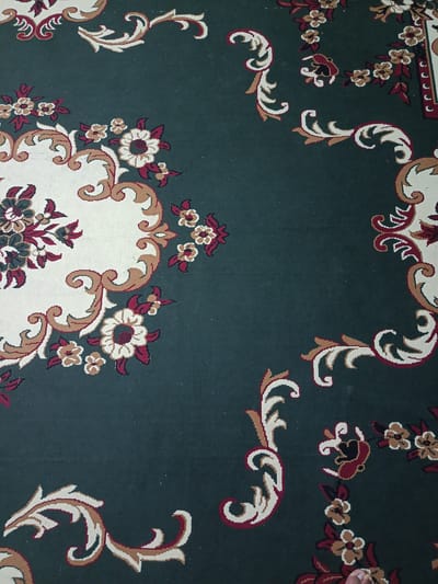 This media file displays a close-up, top-down view of a dark green carpet with an ornate floral and scroll pattern. The design features swirling vines and stylized flowers in cream, burgundy, and brown. Some of the flowers are depicted with multiple petals, while others are simpler, four-petaled shapes. The patterns are intricate and repeat across the carpet. The lighting suggests an indoor setting, likely with ambient light. No people are clearly visible, though a small portion of a bare foot is visible at the bottom right edge of the frame. The scene is static, focusing solely on the carpet's design. There is no visible text. The location context is Ӯртабуз, Tajikistan, but this information cannot be inferred from the visual content of the image itself.