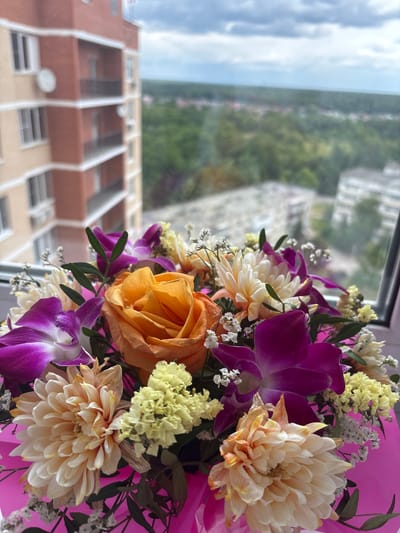 The image shows a close-up view of a vibrant bouquet of flowers, including orange roses, purple orchids, and creamy white and yellow chrysanthemums, arranged in a pink wrapper. The bouquet is positioned in front of a window, with a blurred background revealing a residential building with a brick and beige facade. Beyond the building, a landscape of green trees and other structures can be seen, under a cloudy sky. The context of Dubovaya Roshcha, Russia is implied by the question, suggesting the building and surrounding landscape are characteristic of that location. No people are visible in the image.
