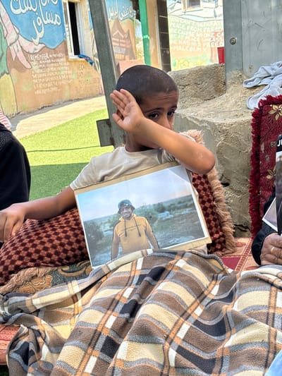 This image depicts an outdoor scene in Carmel, Palestinian Territory, centered on a young boy. The boy, appearing pre-teen with short dark hair, wears a light-colored t-shirt. He is seated on a brown and red patterned cushion, partially covered by a brown, tan, black, and orange plaid blanket that also extends over the lap of an unseen individual to his right. He holds a framed photograph of a smiling man in a yellow hoodie and a dark bucket hat, set against a natural outdoor backdrop. The boy's left hand is raised, partially covering his face, and his eyes appear sad. To the boy's immediate right, an adult's hand with a ring is visible resting on a red and gold patterned mat beneath the plaid blanket. In the background, a light-colored building wall is adorned with a colorful mural and text in both English and Arabic. Visible English phrases include "I REMEMBER MY MOTHER'S FACE...", "WE CAN FEEL SAD BUT WE CAN'T BE DEMORALIZED...", and "YOU CAN'T SEPARATE PEACE FROM FREEDOM BECAUSE NO ONE CAN BE AT PEACE UNLESS HE HAS HIS FREEDOM MALCOLM X". The ground directly in front of the wall appears to be green artificial turf, while a concrete or stone structure is visible behind the boy. The scene is illuminated by daylight.