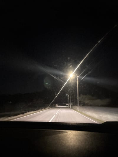 The image depicts a nighttime scene viewed from the interior of a vehicle, looking forward through a windshield. The windshield exhibits multiple streaks and circular splatters, along with a distinct bright light source creating lens flares, including a prominent green circular artifact in the upper left-center. A two-lane asphalt road, marked with solid white lines along the shoulders and a dashed white center line, extends into the distance, illuminated primarily by overhead streetlights. Several utility poles, some supporting operational streetlights, line the right side of the road. The immediate foreground shows the vehicle's dashboard, with reflections of ambient light. The environment beyond the road is dark, indicating night, with vague, dark shapes suggesting distant hills or dense vegetation. No people are visible. This scene is located in Carrazeda de Ansiães, Portugal.