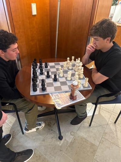 This image depicts an indoor scene within a common area in Waltham, United States, where two young adult males are engaged in a game of chess at a round, dark wood-grain table.
The subject on the left, a male with dark curly hair, wears a black hoodie, olive green cargo pants, and white sneakers with a black star design. He is seated, looking intently at the chessboard. The subject on the right, a male with wavy brown hair, wears a black t-shirt, olive green cargo pants, and dark slip-on shoes. He is seated, with his right hand raised near his mouth, indicating contemplation while observing the chessboard. Partially visible in the lower left corner are the legs and feet of a third individual, wearing black pants and dark slip-on shoes.
The chessboard on the table features black and white plastic chess pieces, arranged mid-game. A brown plastic bottle with a white cap, likely chocolate milk, rests on a white paper sheet (possibly a menu or flyer) near the right player. The table is supported by black metal legs. The chairs are black metal-framed, with a visible blue seat cushion on the right chair. The background consists of light brown wooden wall paneling. A vertical silver-colored pole extends from the floor to the ceiling on the right side of the background. A white rectangular device is mounted on the wall to the left of the pole. The floor is covered with light-colored, square tiles, displaying several circular stains or marks.