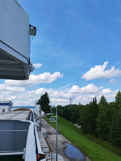 This image depicts a section of the Belousovo Hydroelectric Power Plant, located in the city of Belousovo, Russia. In the foreground, a large white structure, likely part of a ship or barge, dominates the left side of the frame. Beyond this, a concrete pathway runs alongside a green, grassy embankment. A lamppost with a sign displaying the number "8" is visible on this pathway. In the background, a dense forest of green trees stretches across the landscape under a bright blue sky dotted with white clouds. Several buildings, including what appear to be control towers, are visible on the left side of the image, near a body of water. Electrical transmission towers can be seen in the distance, amidst the treeline. The scene is bathed in daylight, suggesting it was taken on a clear day.