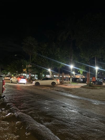 This image depicts a nighttime street scene in Camaragibe, Brazil. The environment is dark, illuminated by multiple overhead streetlights and the headlights and taillights of vehicles. The road surface in the foreground and midground is wet, heavily puddled, and appears to be uneven or damaged.
On the far left, the rear bumper and tailgate of a white pickup truck are partially visible. Further along the road, a white SUV with illuminated taillights is positioned on the left, followed by another vehicle obscured by the SUV's light. A white sedan is prominently centered in the midground, facing away from the camera, seemingly stopped or moving slowly. This sedan is positioned near an open gate or entrance on the right, which leads into a dirt or gravel parking area.
The parking area contains several dark-colored vehicles parked irregularly. A single individual, wearing an orange high-visibility vest, stands on the far right side of this parking area near a parked car.
Numerous trees, including visible palm trees, line both sides of the street, their foliage illuminated by the streetlights. In the background, dimly lit buildings are visible, and street signs are discernible on poles, with one sign clearly displaying the word "GESTO." Reflections from the bright streetlights are prominent on the wet road surface.