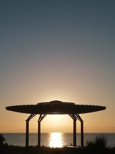 An outdoor, wide-angle shot captures the sunset over the Issyk-Kul lake near Kaji-Say, Kyrgyzstan. A dark, silhouetted structure resembling a UFO with four supporting pillars stands in the foreground, positioned over the calm, shimmering water. The sun, a bright orb, is visible behind the structure, casting a golden glow on the water's surface. The sky transitions from a light blue at the top to a soft orange hue near the horizon. The lake is bordered by a thin strip of land with dark vegetation in the foreground and faint, mountainous silhouettes in the background.