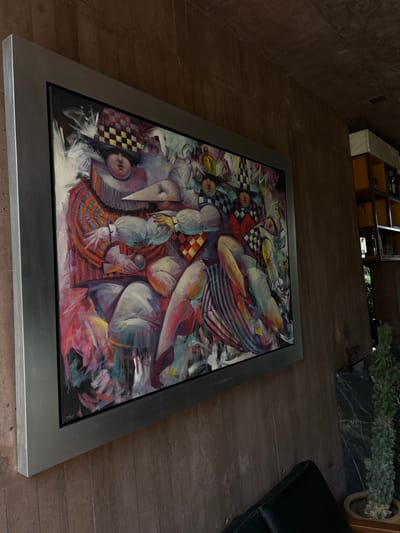 The image shows a framed painting lying on a textured floor, likely stone or concrete. The painting features an abstract composition with figures resembling harlequins or clowns, rendered in a painterly style with vibrant colors and checkerboard patterns. A black leather sofa is partially visible on the right side of the frame. In the background, there is a built-in shelving unit with various items displayed on it. Additionally, a potted plant sits atop a dark-colored plinth near the shelves. Given the artistic style and architectural features, the location is likely within a private residence or gallery in Tepoztlán, Mexico.