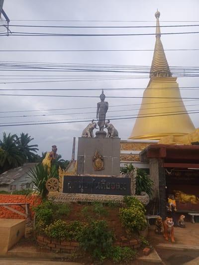The image shows a Buddhist temple complex in Ban Thung, Thailand. A golden stupa dominates the background. A statue of Buddha stands on a pedestal flanked by elephant sculptures. A sign with Thai script sits in front of the monument, surrounded by foliage. Statues of tigers and other figures are present near a small structure to the right. Power lines are visible in the sky above. Palm trees and a residential building appear in the background. The environment suggests a partly cloudy day.