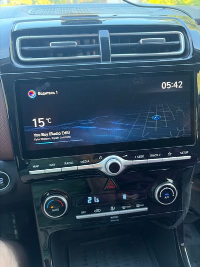 The image shows the interior of a modern car's dashboard, specifically the infotainment screen and climate controls. The infotainment screen displays the time as 05:42, the current temperature as 15°C, and the currently playing song as "You Boy (Radio Edit)" by Kyle Watson and Kylah Jasmine. It also shows a navigation map with a blue marker indicating a location. Below the screen, there are control buttons for "MAP," "NAV," "RADIO," and "MEDIA." To the right of these buttons are controls for seeking tracks and setting up the system. Further down, the climate control panel features a dial with "AUTO" and indicators for temperature settings, and another dial labeled "A/C" for air conditioning. There is also a digital display showing "21.5" possibly representing a temperature setting, and buttons for "OFF," "FRONT," and "REAR" defrosters. The dashboard is glossy black with metallic accents. The environment suggests the car is parked, likely in a well-lit area given the brightness of the screen. No people are visible in the image. The location is stated to be Skopin, Russia, though the image itself does not contain any direct visual indicators of this specific location.