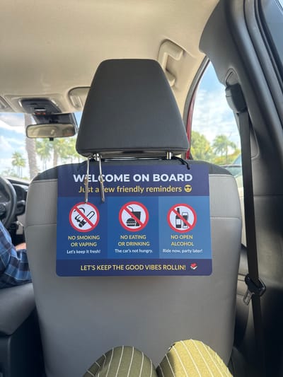 A digital photo shows the interior of a car. The photo is oriented vertically. A blue sign is hanging from the headrest of a car seat. The sign reads, "WELCOME ON BOARD Just a few friendly reminders". It also lists rules: "NO SMOKING OR VAPING Let's keep it fresh!", "NO EATING OR DRINKING The car's not hungry.", and "NO OPEN ALCOHOL Ride now, party later!". The sign concludes with "LET'S KEEP THE GOOD VIBES ROLLIN!". The car appears to be parked outdoors, as indicated by the sky and palm trees visible through the car's windows. The weather seems to be sunny. The photo captures a glimpse of the car's steering wheel and a person's shoulder in a plaid shirt, suggesting a passenger or driver is present. The scene is casual and friendly, emphasizing a positive experience for the car's occupants. The location is identified as Hallandale Beach, USA. The sign indicates an event or activity related to shared transportation, likely a ride-sharing service or a shared carpool, where basic courtesies are being outlined for the passengers. The overall tone is lighthearted and aims to ensure a pleasant and clean environment for everyone. The image is taken from the perspective of someone sitting in the back of the car, looking forward. The headrest and part of the car seat are the most prominent objects in the foreground, with the sign being the central focus. The background elements, such as the sky and foliage, provide context about the outdoor setting. The lighting suggests it is daytime, possibly midday, given the brightness of the sunlight. The overall mood is cheerful and welcoming. The text on the sign is a key element, providing specific instructions and setting expectations for the passengers. The use of emojis on the sign further enhances the friendly and informal tone. The image is a snapshot of a common experience in modern transportation, highlighting the communication of rules and expectations in a visually engaging way. The photograph is taken from a slightly elevated position within the car, looking towards the front headrest. The angle highlights the sign and its message. The details of the car's interior, like the seat fabric and the visible portion of the dashboard, are secondary to the sign's content. The location context of Hallandale Beach, USA, suggests a potentially warm and sunny climate, which aligns with the visual cues of the bright sky and greenery. This