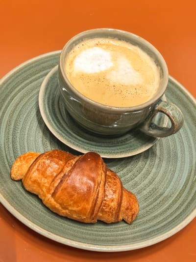 A close-up, high-angle shot shows a croissant and a cup of coffee with latte art on a green plate. The croissant is golden brown and flaky, positioned on the left side of the plate. The coffee, served in a textured gray mug, is in the upper right quadrant of the image, with foamy milk art on top. The plate has a ribbed texture and a light-colored rim, and it sits on an orange surface, likely a table. The background is a solid, vibrant orange. The lighting suggests it's daytime, possibly indoors in a cafe. The overall impression is one of a simple, pleasant breakfast or snack. There is no visible text.