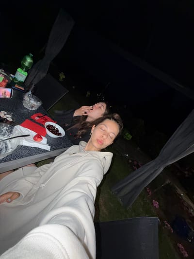 Two young women are at a table outdoors at night. One woman wears a light-colored hoodie and makes a kissing face toward the camera. The other woman eats. A bowl of cherries, a peach, red napkins, empty bowls, a green glass bottle, and a phone sit on the table. The location is Hanau, Germany.