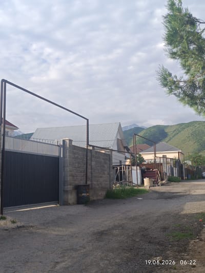 The image depicts a street scene in the city of Қырғауылды, Kazakhstan. A large, dark gray metal gate dominates the left side of the frame, flanked by a textured gray stone wall. Beyond the wall, several houses with varied roofing materials are visible, including corrugated metal and what appears to be asphalt shingles. In the background, lush green mountains rise under a cloudy sky. A large green tree with dense foliage partially obscures the upper right portion of the image. The ground in the foreground is a gravel road, with some sparse grass growing along the edges. A rusted metal barrel is positioned near the gate. The timestamp "19.06.2026 06:22" is visible in the lower right corner. No people or specific actions are clearly discernible.
