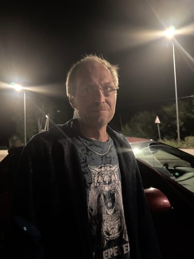 A waist-up, medium shot captures a light-skinned man with short blonde hair and glasses, wearing a dark jacket over a t-shirt with a black and white graphic of a woman with a wolf headdress and another wolf below her. He is standing outdoors at night, with streetlights visible in the background. A red car is parked to his right, partially obscuring the lower right portion of the image. The scene appears to be a street or parking area with trees and a small sign in the distance. The image is taken from a slightly low angle, looking up at the man. The lighting is dramatic, with strong highlights from the streetlights creating a lens flare effect.